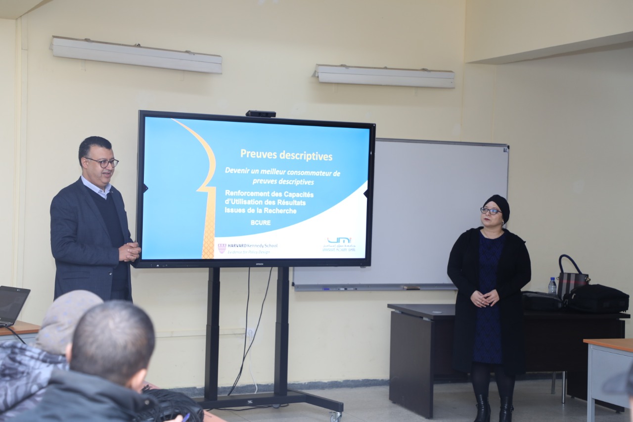 One male and one female Moroccan professor stand in front of a presentation screen in a classroom. The screen has text in French that translates to "Descriptive Evidence: Becoming an Effective Consumer of Descriptive Evidence."